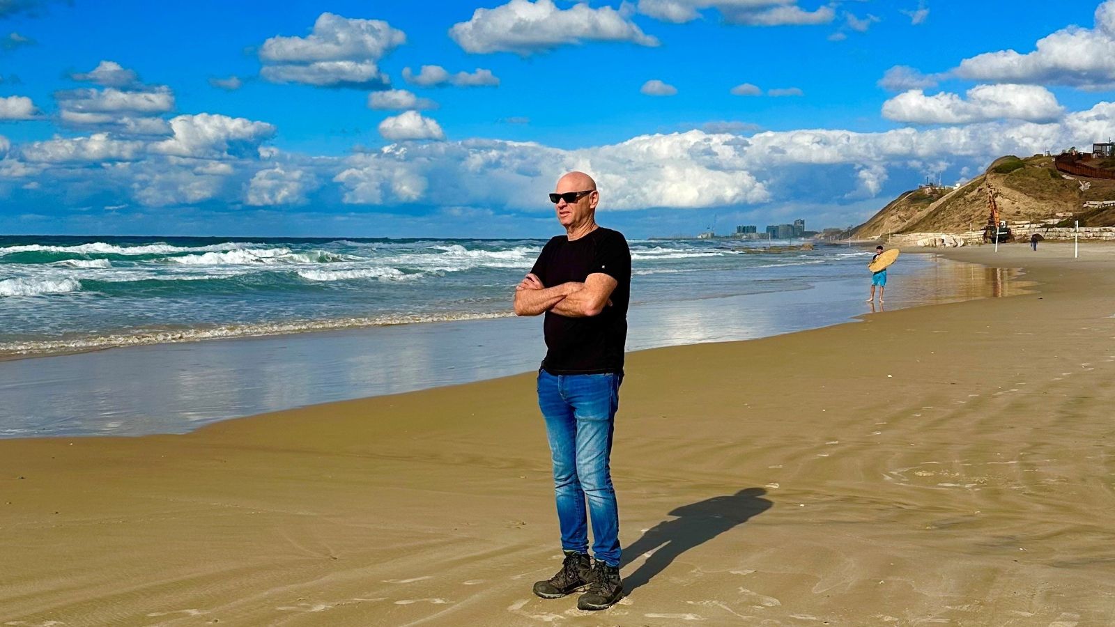 A man in a black t-shirt and blue jeans stands on a beach. His arms are crossed and he is looking out at the water.