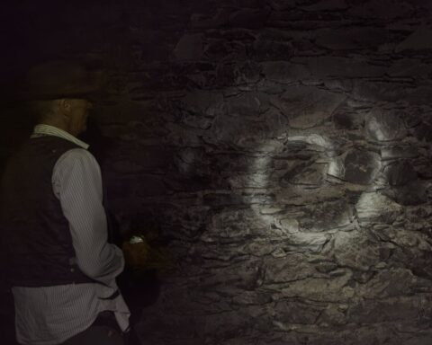 A man in a dark cave stands with his back to the camera, holding a flashlight towards the cave wall while looking at a white spiral on the rocks.