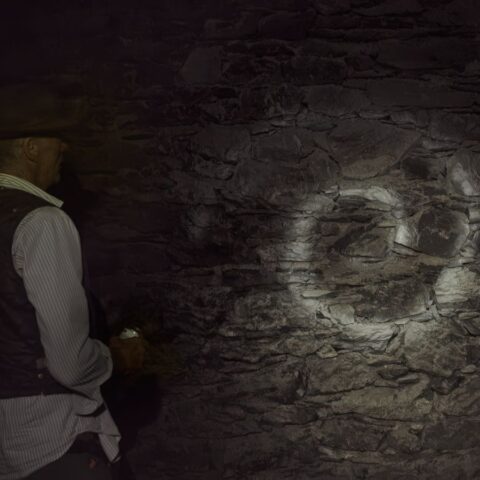 A man in a dark cave stands with his back to the camera, holding a flashlight towards the cave wall while looking at a white spiral on the rocks.