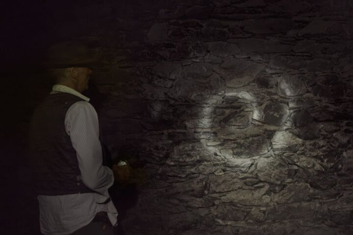 A man in a dark cave stands with his back to the camera, holding a flashlight towards the cave wall while looking at a white spiral on the rocks.