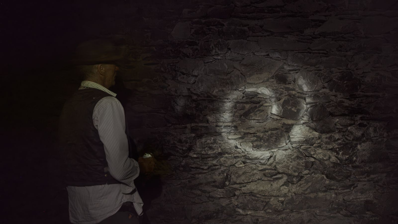 A man in a dark cave stands with his back to the camera, holding a flashlight towards the cave wall while looking at a white spiral on the rocks.