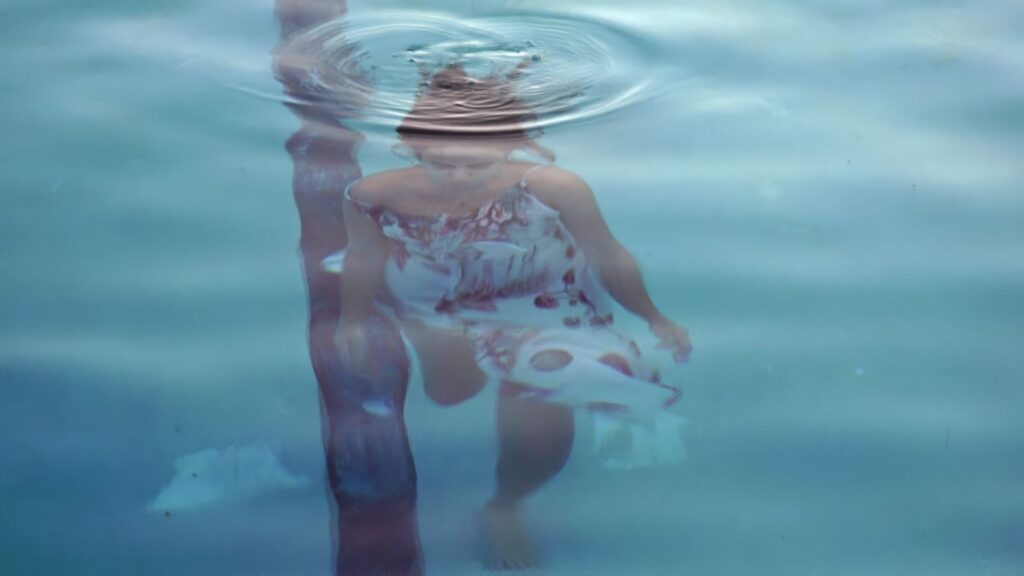 A view looking down upon a woman who is below the surface of the water. There is a ripple above her. She has brown hair and is wearing a white sun dress.