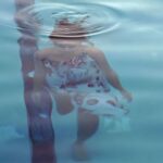 A view looking down upon a woman who is below the surface of the water. There is a ripple above her. She has brown hair and is wearing a white sun dress.