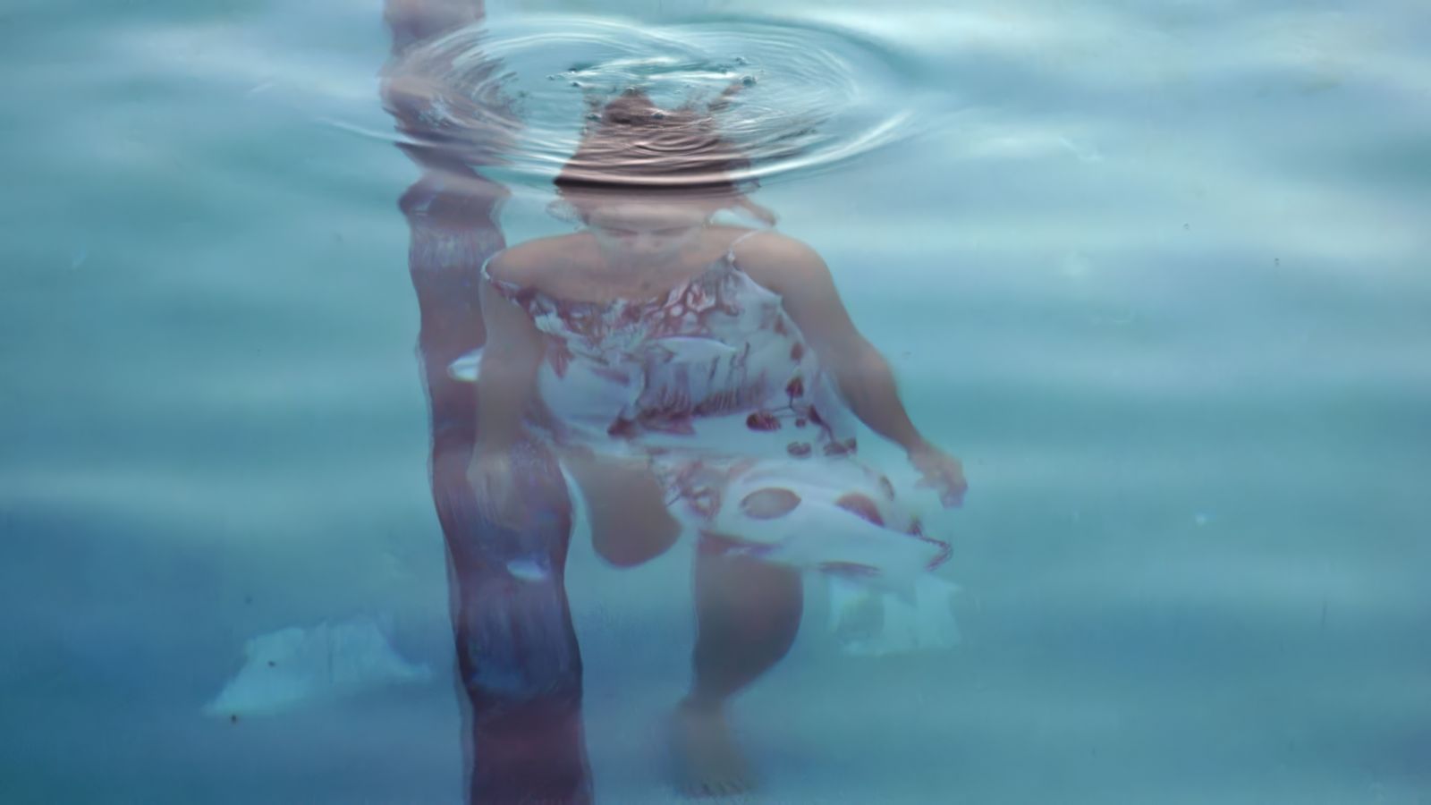 A view looking down upon a woman who is below the surface of the water. There is a ripple above her. She has brown hair and is wearing a white sun dress.