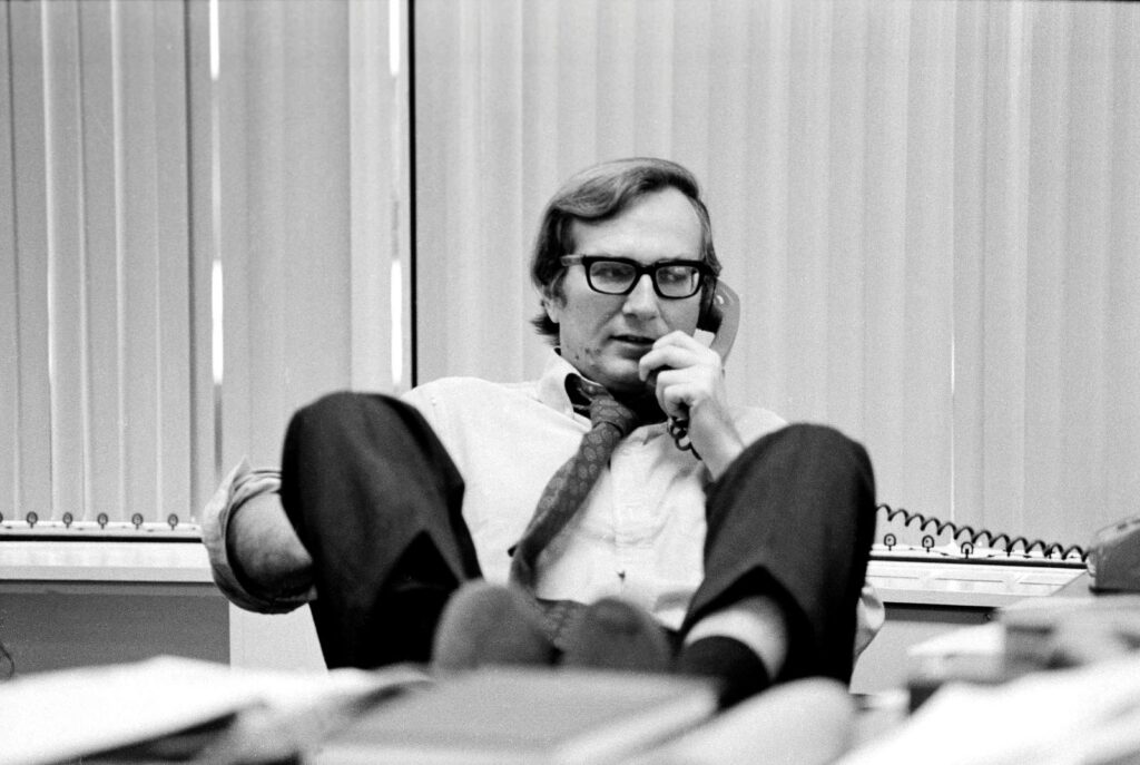 A black and white photo of journalist Seymour Hersh. He is seated at his desk and talking on the telephone with his feet propped on the edge.