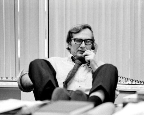 A black and white photo of journalist Seymour Hersh. He is seated at his desk and talking on the telephone with his feet propped on the edge.