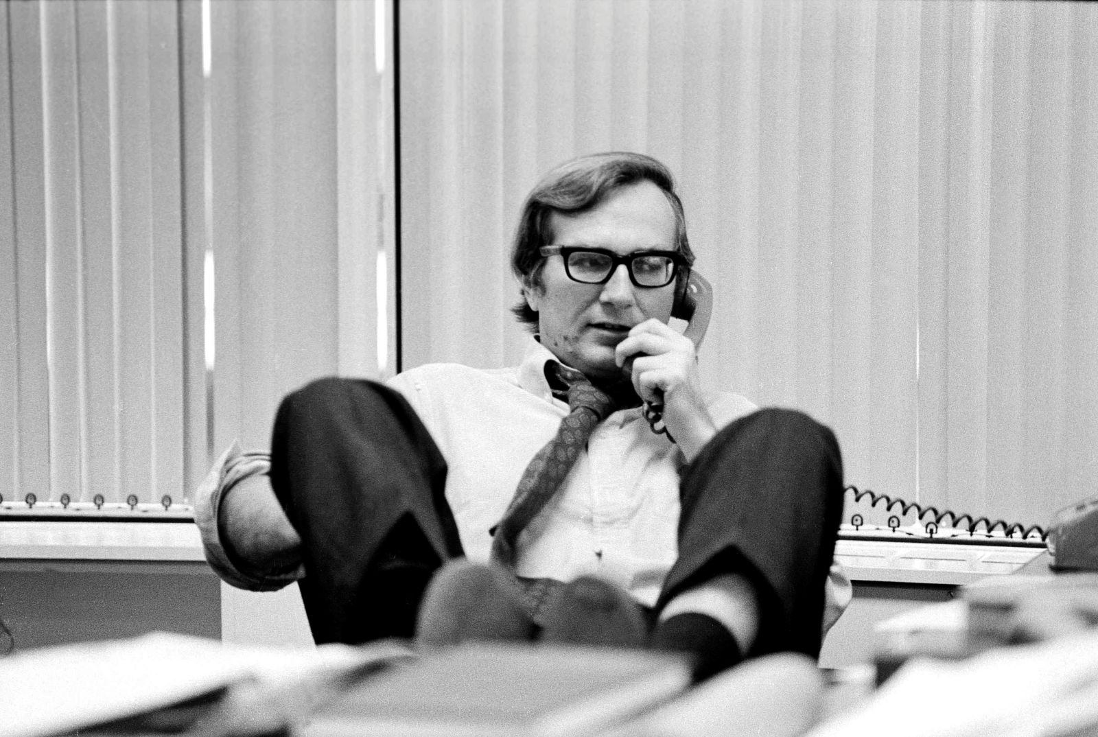 A black and white photo of journalist Seymour Hersh. He is seated at his desk and talking on the telephone with his feet propped on the edge.