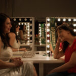 Filmmaker Brishkay Ahmed and participant Vida Samadzai have a discussion in a dressing room. They are sitting in front of two mirrors framed with light bulbs.