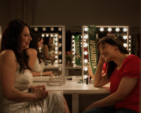 Filmmaker Brishkay Ahmed and participant Vida Samadzai have a discussion in a dressing room. They are sitting in front of two mirrors framed with light bulbs.