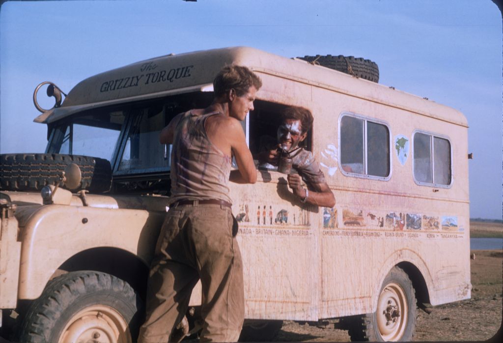 An archival 16mm film of two friends in 1957 with their Land Rover. One is seated in the driver's seat, while the other stands in front of the beige camper van.