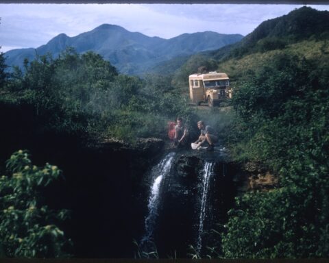 An archival 16mm film still of two friends sitting at the top of a waterfall. Their beige Land Rover is in the distance.
