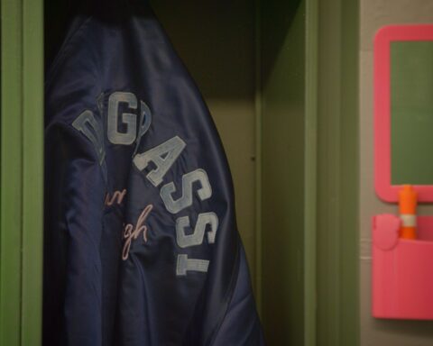 A school jacket hangs in a closet. It is blue with the letters DEGRASSI in white.