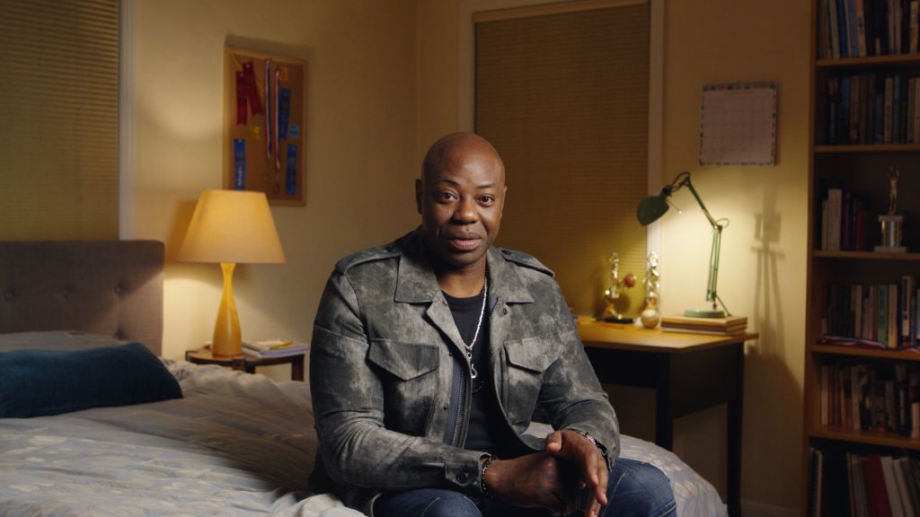 Actor Dayo Ade sits on a bed while giving an interview.