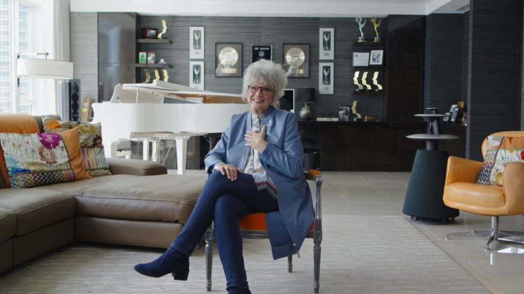 Degrassi creator Linda Schuyler sits in a living room while giving an interview.
