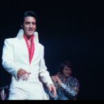 An archival image of Elvis Presley on stage in his white coverall suit with a red shirt.