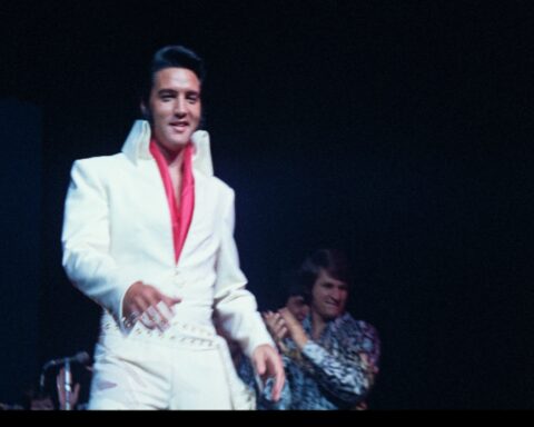 An archival image of Elvis Presley on stage in his white coverall suit with a red shirt.