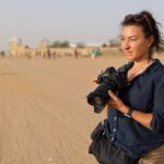 Photographer Lynsey Addario stands in a desert and holds a camera.