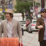 Two 30-something white guys wearing blazers and hats walk down Queen Street West in Toronto with a spool of orange cable.