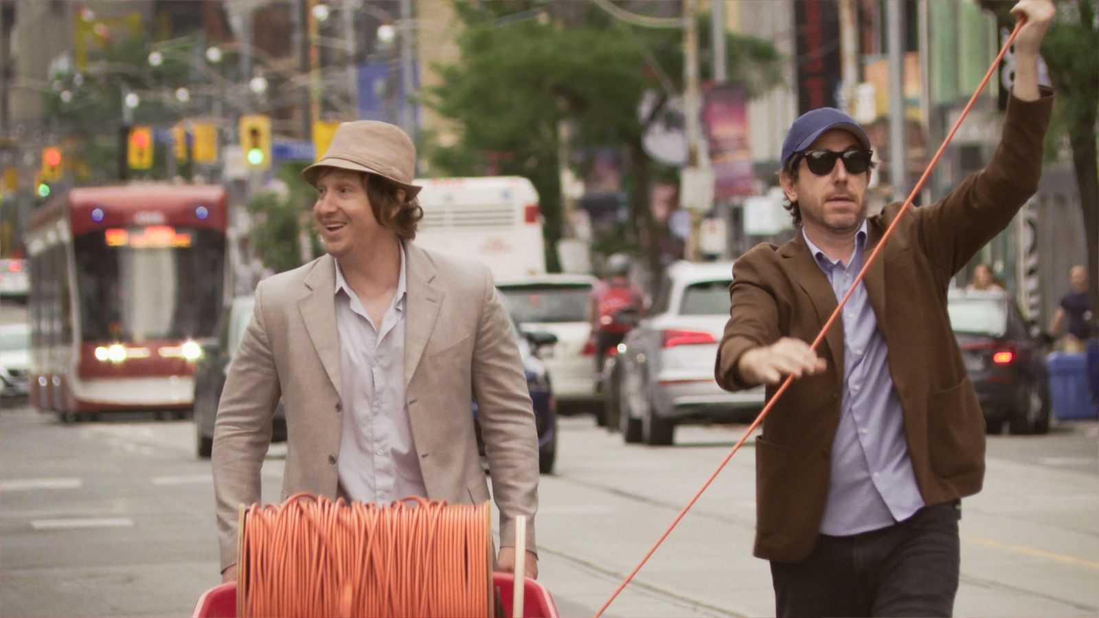 Two 30-something white guys wearing blazers and hats walk down Queen Street West in Toronto with a spool of orange cable.