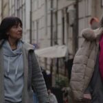 Two women in puffy coats walk through a European street, looking up to the right of the screen.
