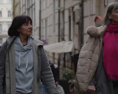 Two women in puffy coats walk through a European street, looking up to the right of the screen.