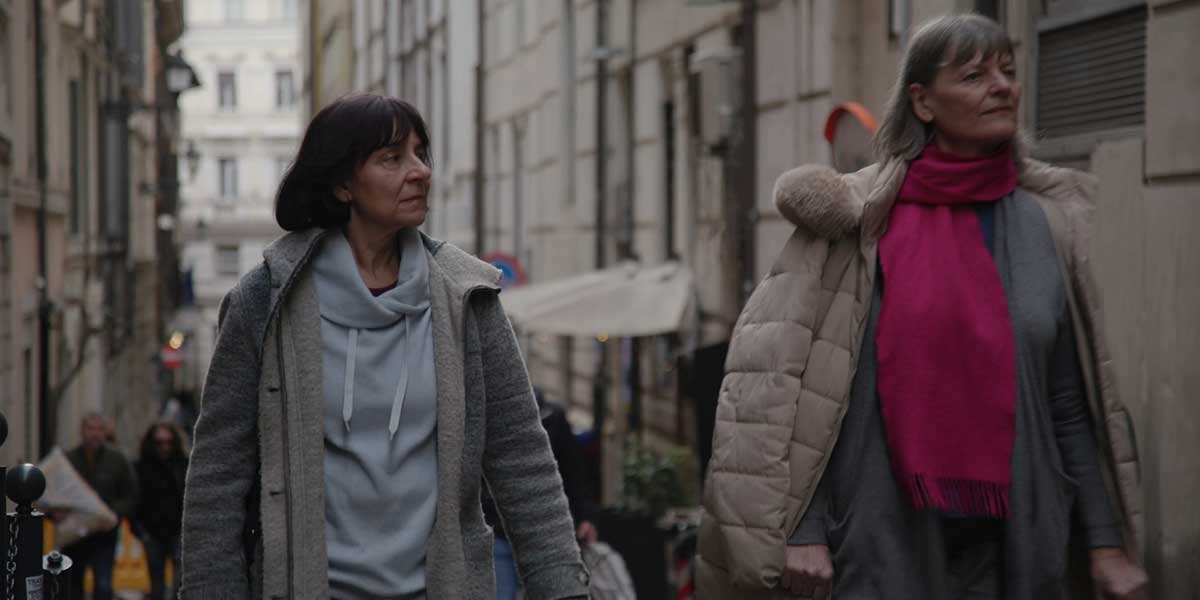 Two women in puffy coats walk through a European street, looking up to the right of the screen.