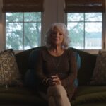 Paula Deen is seated for an interview. She is a white woman with grey hair. Her hands are clasped on her knees, and she is sitting on a couch in her living room.