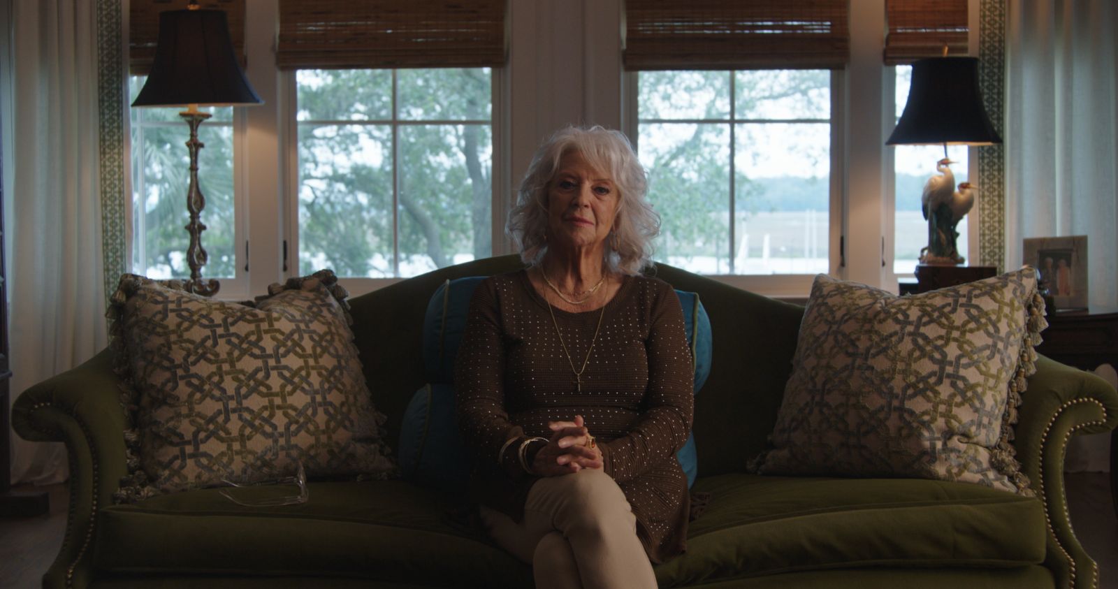 Paula Deen is seated for an interview. She is a white woman with grey hair. Her hands are clasped on her knees, and she is sitting on a couch in her living room.