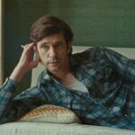 Actor Ben Whishaw lies on a beige couch with his arm propped on his elbow, resting his head on his hand while holding a cigarette. He is a white man with brown hair, wearing a blue plaid shirt.