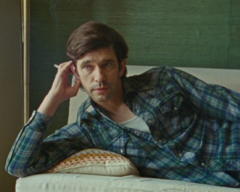 Actor Ben Whishaw lies on a beige couch with his arm propped on his elbow, resting his head on his hand while holding a cigarette. He is a white man with brown hair, wearing a blue plaid shirt.