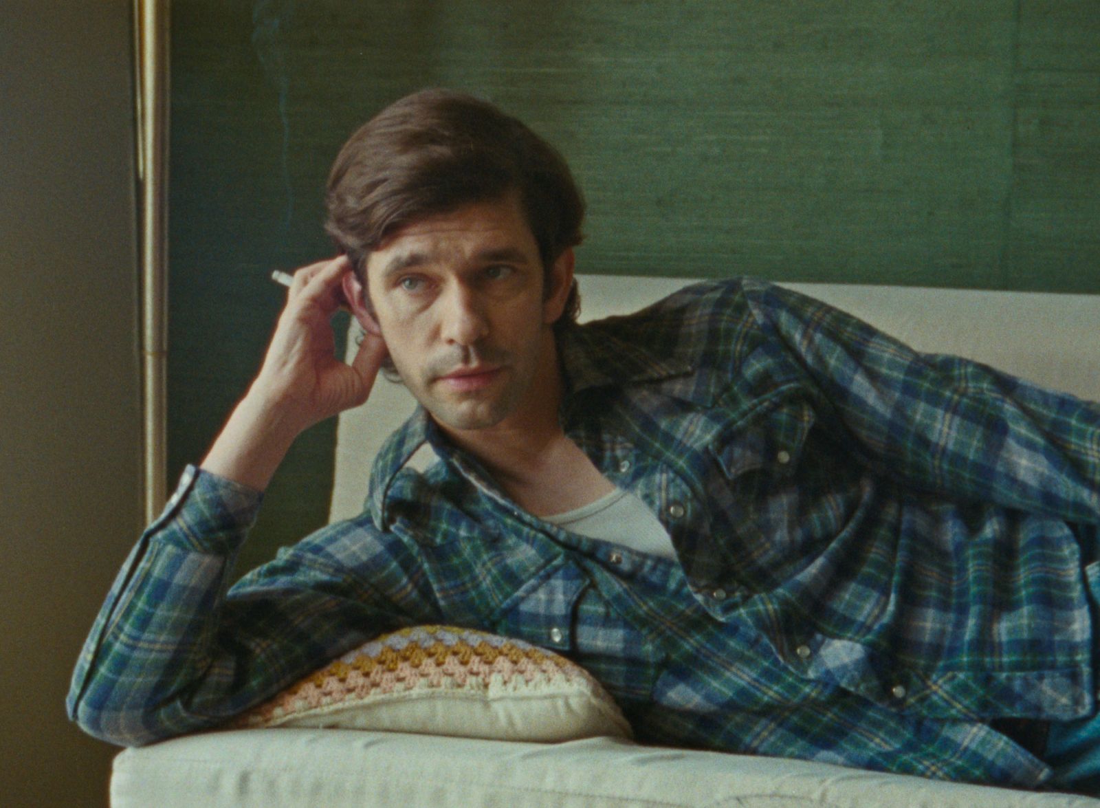 Actor Ben Whishaw lies on a beige couch with his arm propped on his elbow, resting his head on his hand while holding a cigarette. He is a white man with brown hair, wearing a blue plaid shirt.