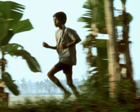 A young South Asian boy runs down the street. He is facing screen left. There are palm trees behind him.