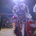 An Indigenous man in full powwow regalia dances before a crowd.