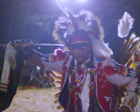 An Indigenous man in full powwow regalia dances before a crowd.