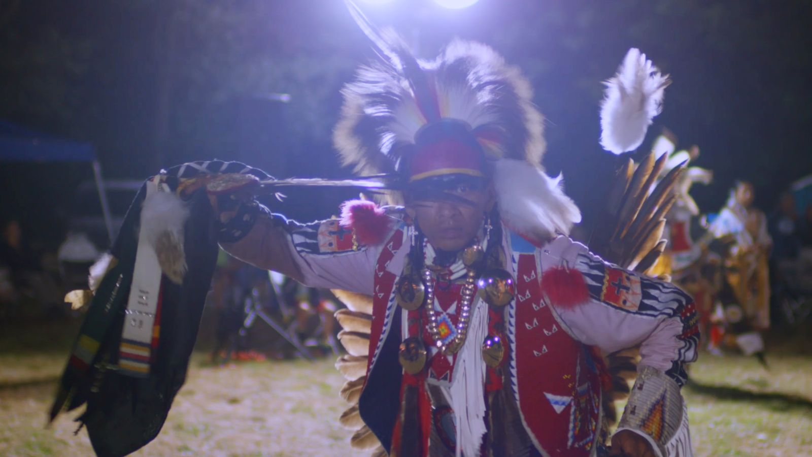 An Indigenous man in full powwow regalia dances before a crowd.