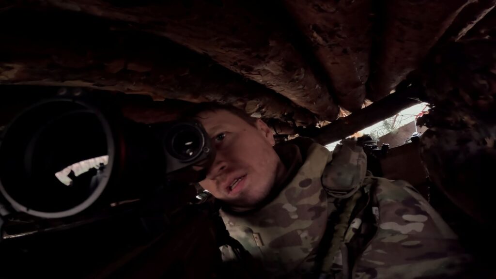 A close up view of a Ukrainian solider under cover and looking through the scope of a sniper rifle.