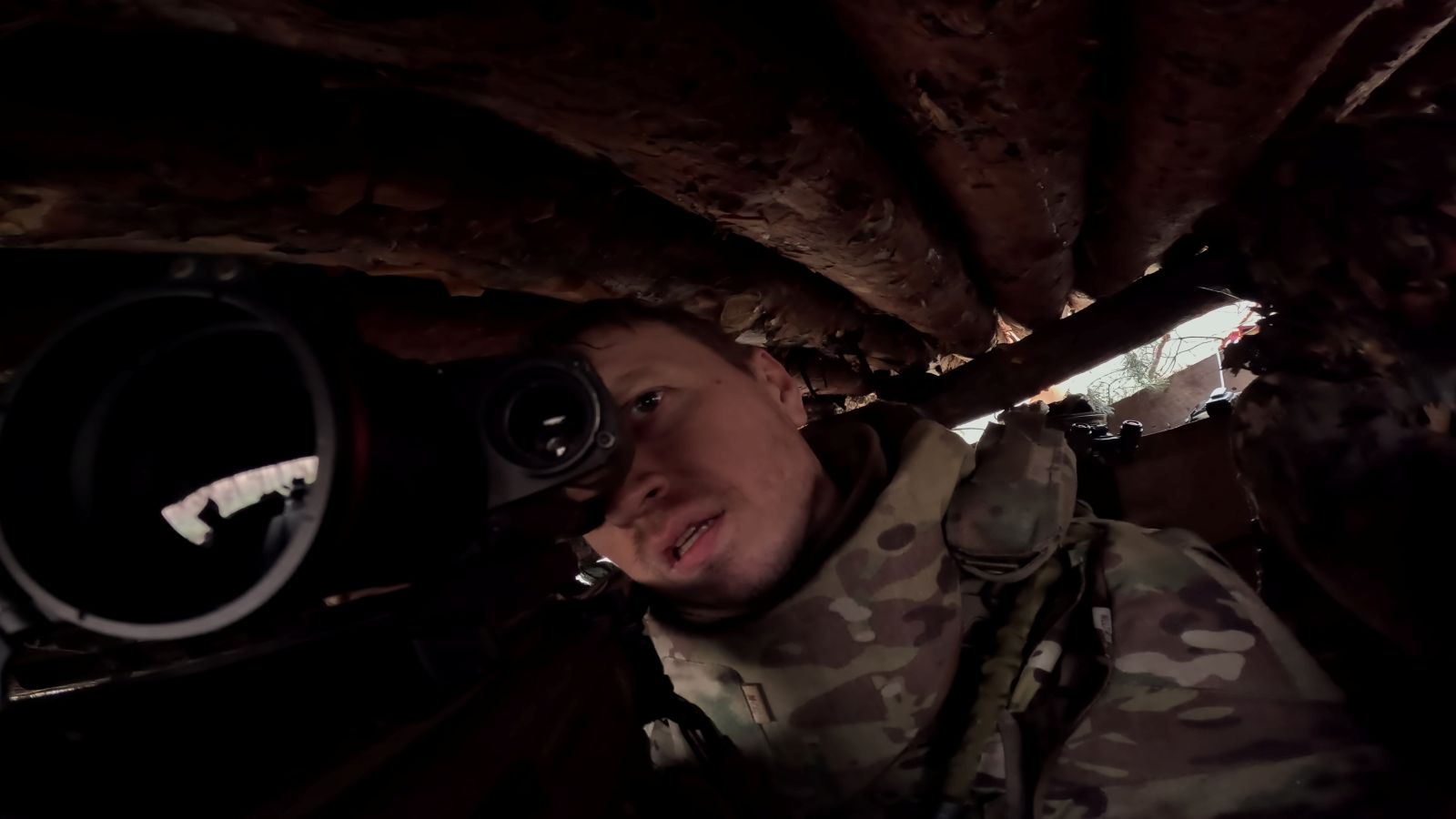 A close up view of a Ukrainian solider under cover and looking through the scope of a sniper rifle.