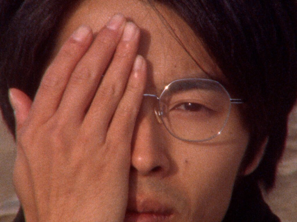 A Japanese man is seen in close up. He holds one hand up to his face covering his right eye.