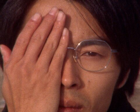 A Japanese man is seen in close up. He holds one hand up to his face covering his right eye.