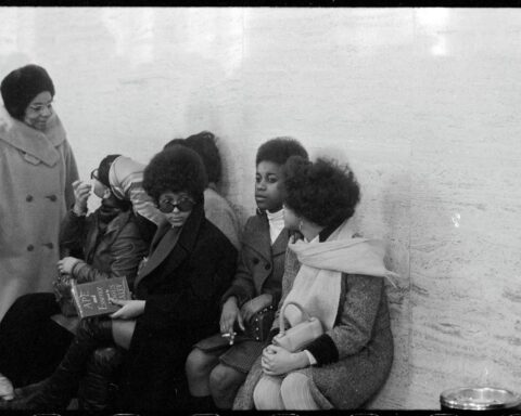 A black and white archival photo of a group of Black women in the 1960s, sitting against a white brick wall and talking.