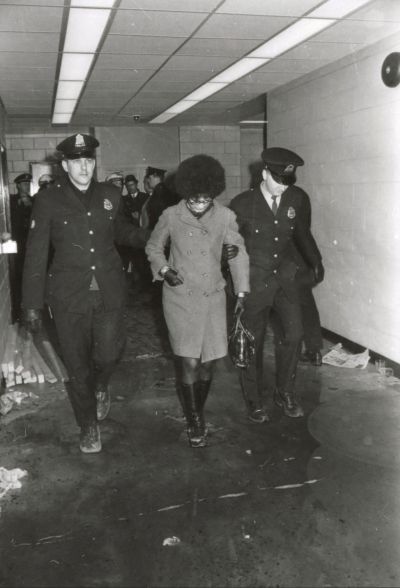 A black and white archival photo of activist Brenda Dash being arrested. She is a Black woman with an afro and overcoat being escorted by two white police officers in uniform.