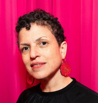 Director Michele Stephenson is a Black Haitian-Canadian woman, pictured in close-up. She is wearing a black shirt, large red earrings, and is pictured against a pink backdrop.
