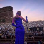 A view behind a woman on stage at a microphone. She is wearing a purple dress and whistling for a crowded audience in an outdoor amphitheatre.