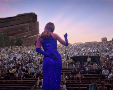 A view behind a woman on stage at a microphone. She is wearing a purple dress and whistling for a crowded audience in an outdoor amphitheatre.