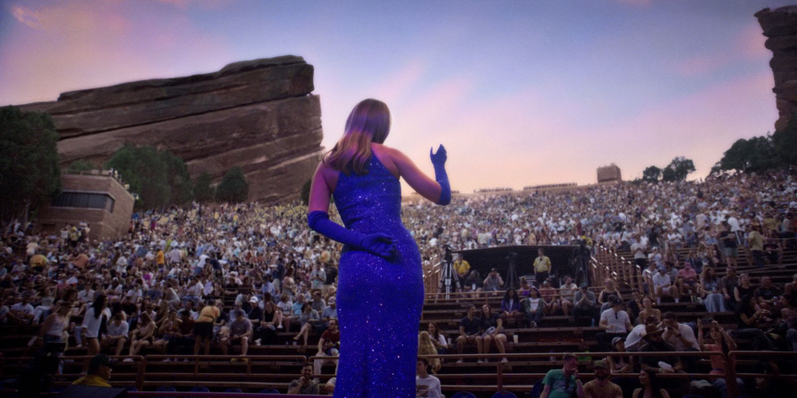 A view behind a woman on stage at a microphone. She is wearing a purple dress and whistling for a crowded audience in an outdoor amphitheatre.