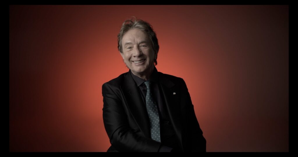 Actor Martin Short is seated for an interview in front of a red background. He is looking at the camera and smiling.