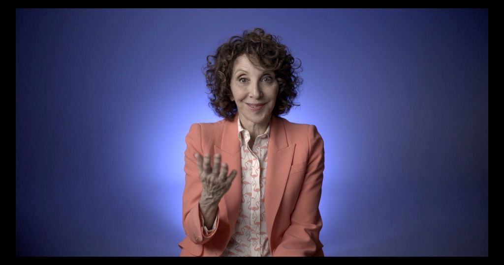 Actor Andrea Martin is seated for an interview, wearing a pink suit with a purple background behind her.