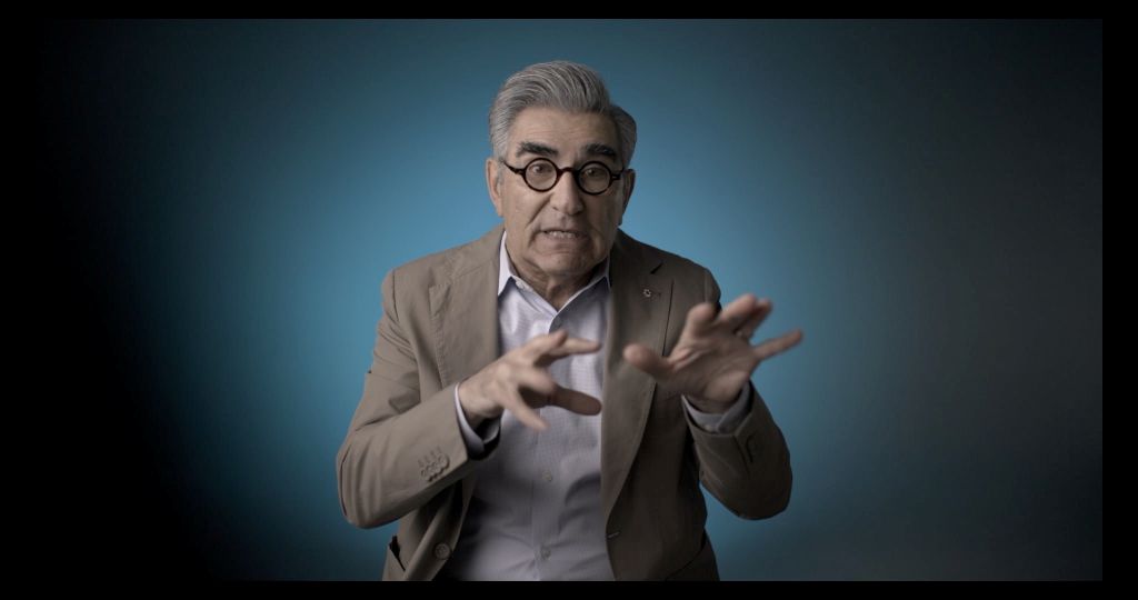 Actor Eugene Levy is seated for an interview. He is wearing a grey suit and has an animated expression with his hands.