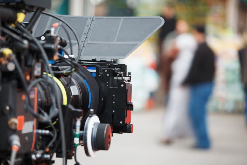 A video camera is in the foreground of the frame and is pointed towards a group of people, who are out of focus in the background.