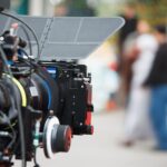 A video camera is in the foreground of the frame and is pointed towards a group of people, who are out of focus in the background.